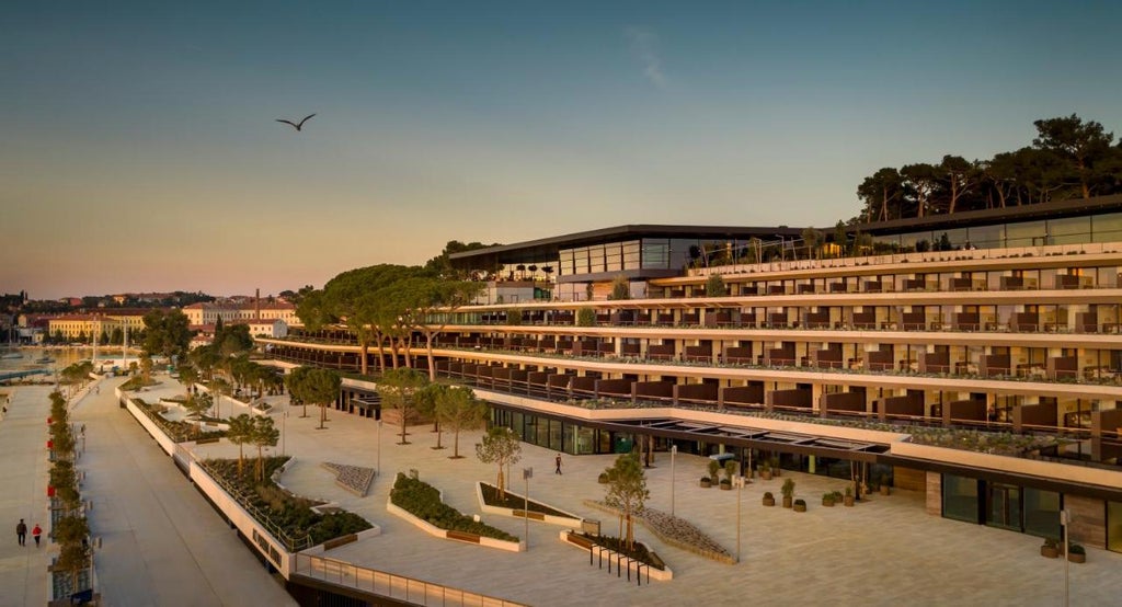 Modern luxury waterfront hotel with curved glass facade, infinity pool overlooking Adriatic Sea, and private marina at sunset.