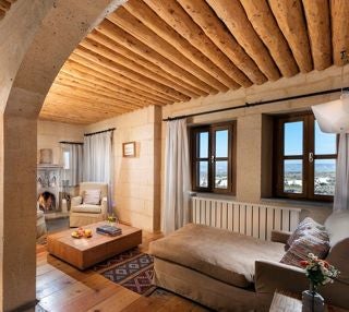 Spacious stone-walled luxury suite with plush white bedding, arched windows overlooking Cappadocia's unique volcanic landscape, blending rustic charm and modern elegance