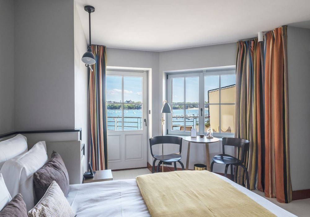 Elegant blue-toned hotel room with panoramic sea view, plush white bedding, modern coastal design, and expansive windows overlooking French coastline