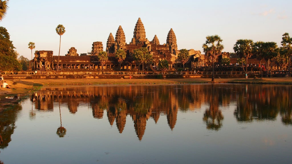 Silhouetted spires of Angkor Wat temple reflect in lotus pond at dawn, with vibrant orange and purple sky illuminating ancient ruins