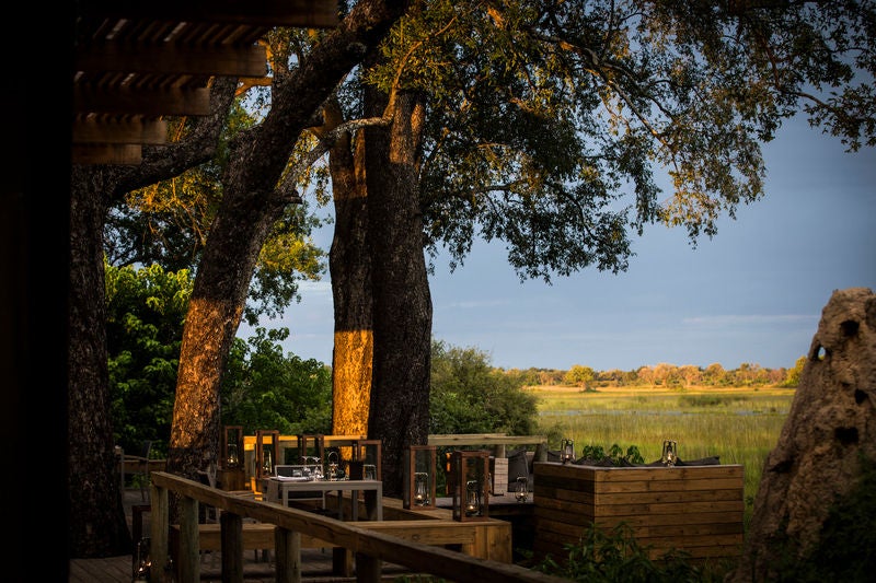 Luxurious safari lodge deck overlooking vast Botswana wetlands, with plush outdoor seating and thatched roof at golden hour