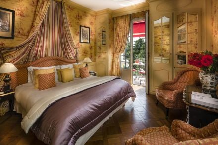 Elegant Provençal-style hotel room with soft blue and white decor, plush white bedding, ornate antique furniture, and large French windows overlooking lush gardens