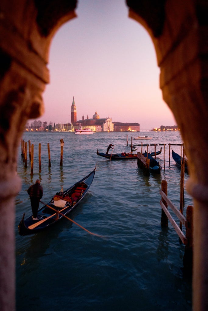 Elegant Venetian palazzo entrance with ornate marble columns, wrought iron gates and traditional gondolas floating on a narrow canal at sunset