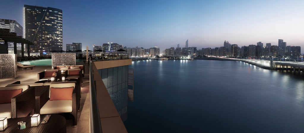 Luxurious waterfront hotel with contemporary glass facade, infinity pool overlooking Arabian Gulf and Abu Dhabi skyline at sunset