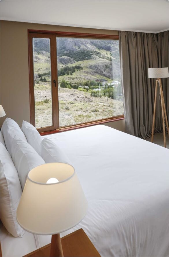 Cozy mountain-view room with wooden floors, plush white bedding, rustic decor, and large windows showcasing breathtaking Argentinian landscape in Los Cerros Boutique Hotel