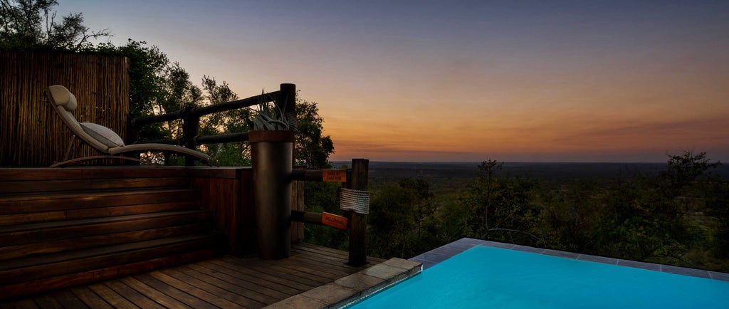 Luxurious stone and wood safari lodge perched atop rocky cliffs in South Africa, overlooking vast savanna with infinity pool at sunset