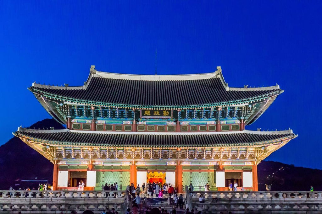 A woman in a vibrant red and blue traditional hanbok dress poses elegantly in front of Gyeongbokgung Palace's ornate wooden architecture and stone walkways.