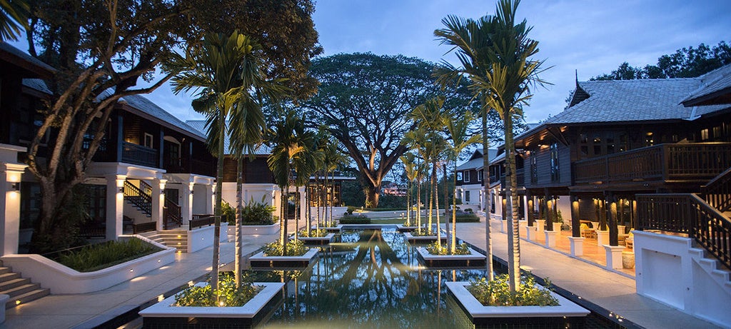 Traditional Thai hotel Na Nirand with wooden balconies and ornate architecture nestled among lush tropical trees beside a tranquil river