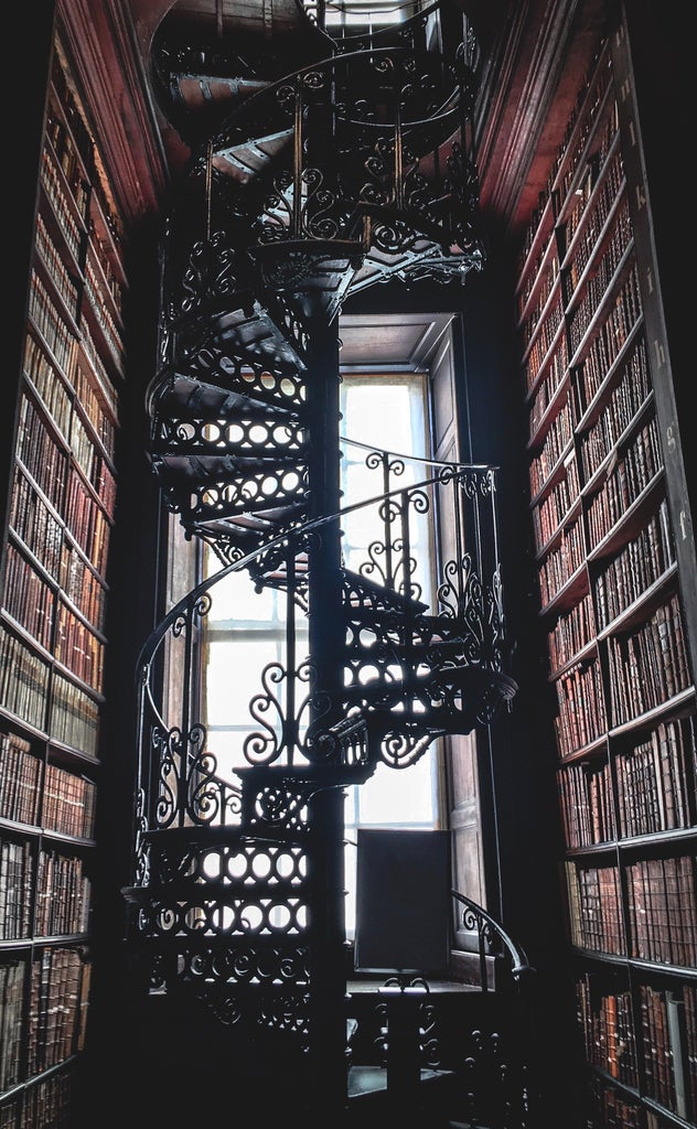 Historic Trinity College library with ornate wooden shelves, grand arched ceiling, and rows of ancient leather-bound books bathed in warm golden light.