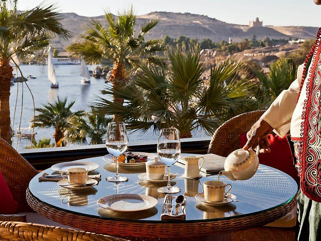 Luxurious Sofitel Legend hotel perched on Nile riverbank, featuring elegant colonial architecture with sandstone walls and lush palm-lined terrace at sunset