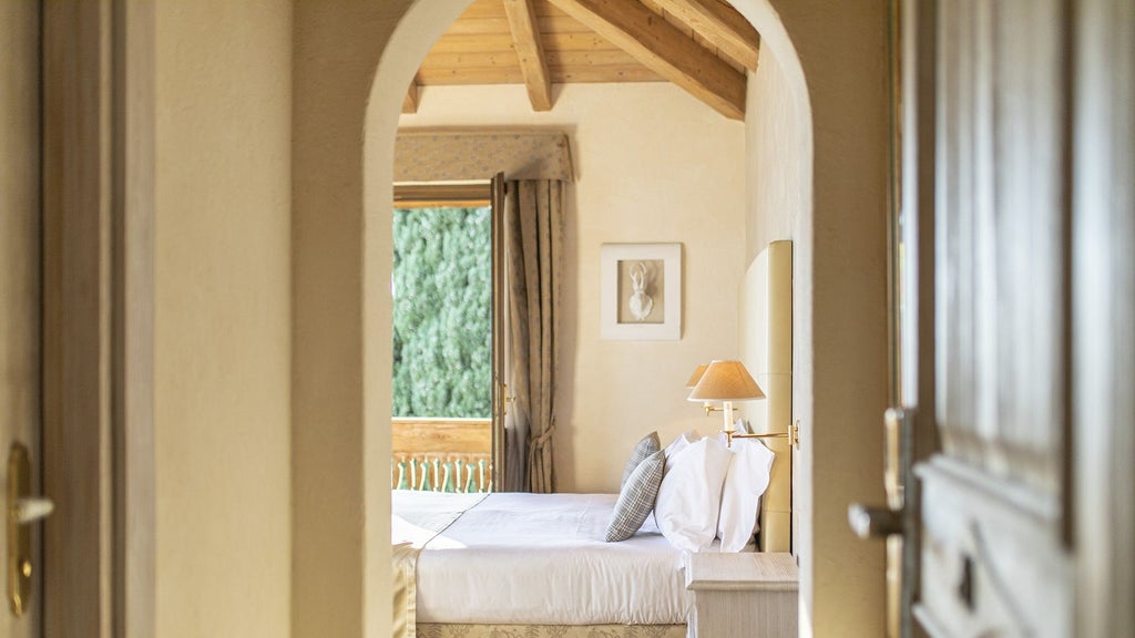 Elegant hotel room with cream walls, plush king bed, ornate headboard, and French doors leading to balcony with partial Lake Garda views