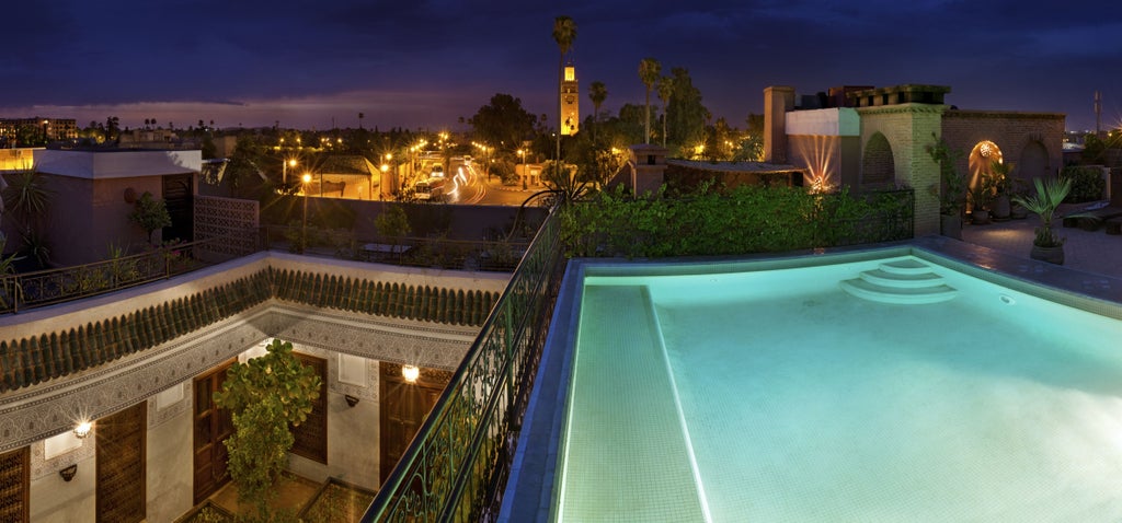 Elegant Moroccan riad with traditional courtyard featuring mosaic fountain, orange trees, and luxurious poolside lounge seating under arched colonnade