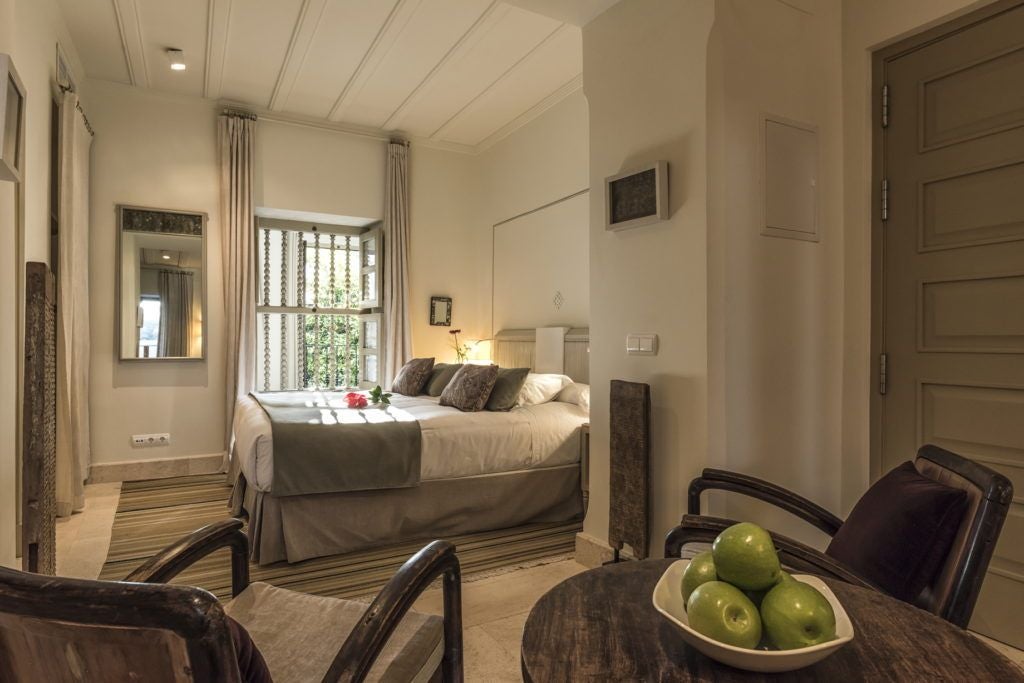 Elegant Spanish-style hotel room with ornate wooden furnishings, white linens, and arched windows overlooking a historic Córdoba courtyard with soft natural light