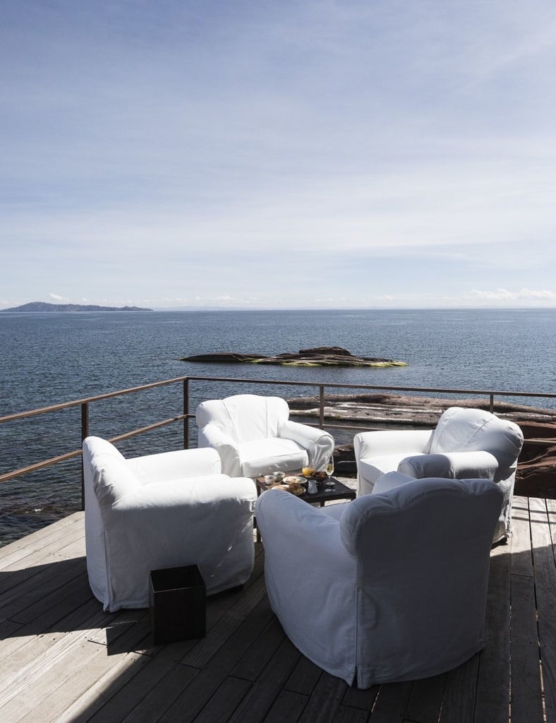 Modern luxury stone lodge with floor-to-ceiling windows overlooking Lake Titicaca, set against dramatic mountain peaks at sunset