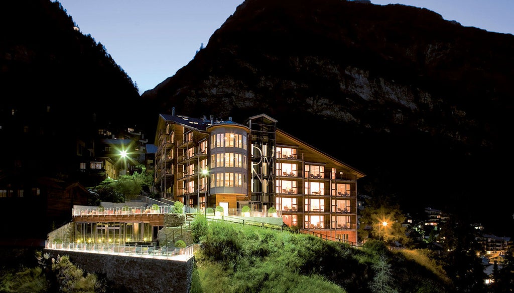 Modern mountain hotel with angular glass-and-wood facade perched on snowy Swiss Alps cliff, illuminated warmly against twilight sky