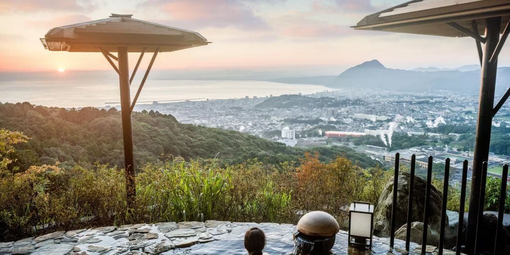 Modern luxury hotel with curved glass facade nestled in mountains, featuring outdoor infinity pools overlooking Beppu city and bay