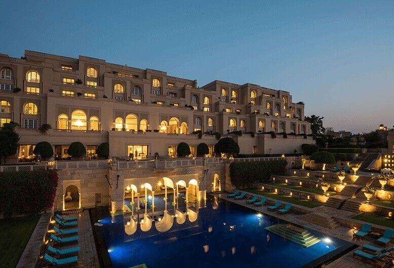 Opulent hotel exterior with Mughal-style domes and archways, overlooking landscaped gardens with fountains, Taj Mahal visible in background