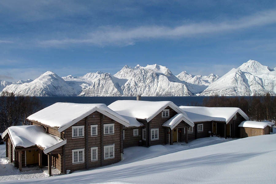 Luxurious wooden lodge nestled in snowy mountains by a fjord, featuring panoramic glass windows and warm exterior lighting at dusk