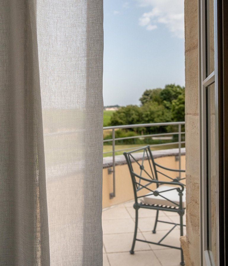 Elegant junior suite with plush white bedding, soft neutral tones, large window overlooking French countryside, classic wooden furnishings, and subtle luxurious design details