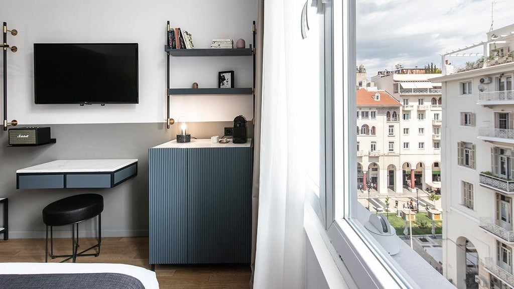 Minimalist hotel room with sleek gray furnishings, white linens, large window, and elegant design showcasing modern Greek hospitality in Thessaloniki