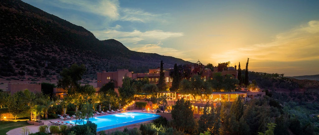 Luxurious Moroccan hilltop hotel with terracotta walls, traditional architecture, and lush gardens overlooking Atlas Mountains at sunset