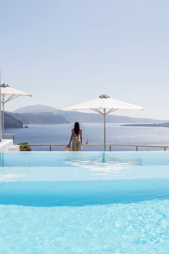 White-washed luxury suites with private infinity pools cascade down Santorini's cliffs, overlooking the deep blue Aegean Sea at sunset