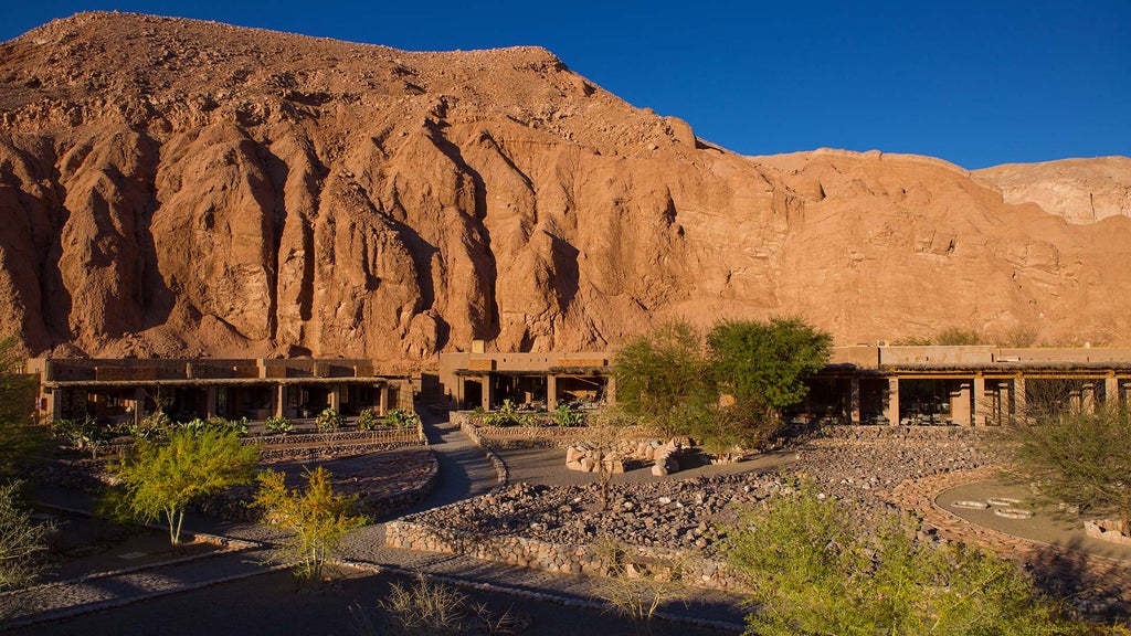 Luxurious desert lodge with adobe-style buildings nestled against red mountains, featuring an infinity pool at sunset in Atacama