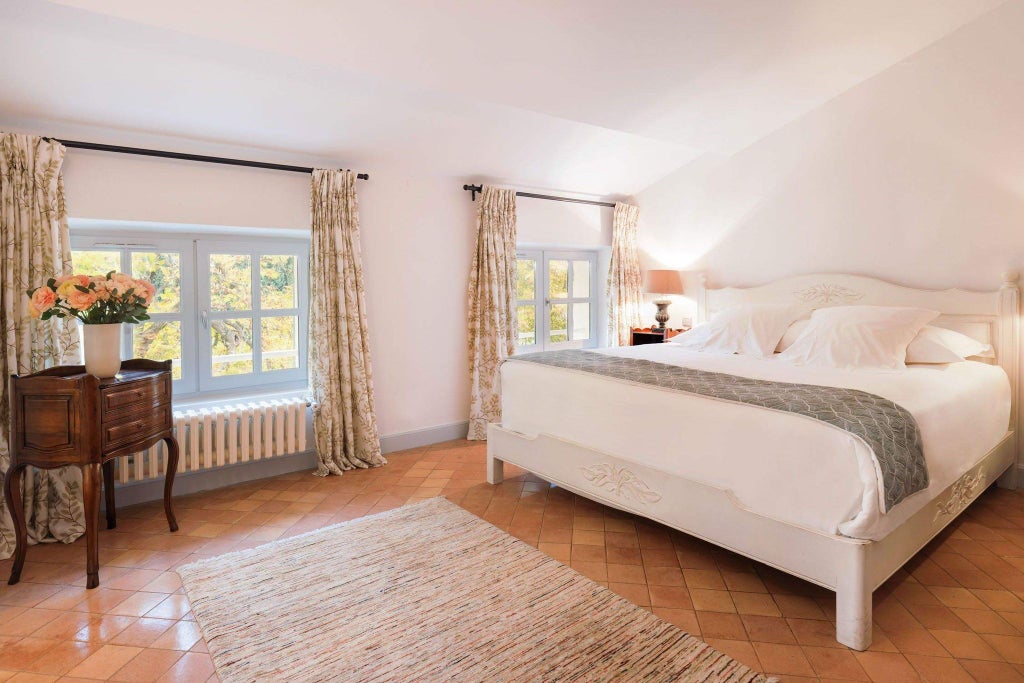 Elegant French countryside hotel room with soft neutral tones, antique furnishings, and large arched window overlooking lush garden landscape