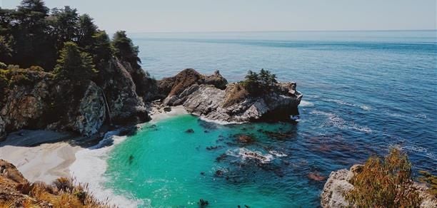 Luxurious villa suite with panoramic coastal views, rustic wood furnishings, king bed, and expansive windows overlooking scenic Big Sur landscape at sunset