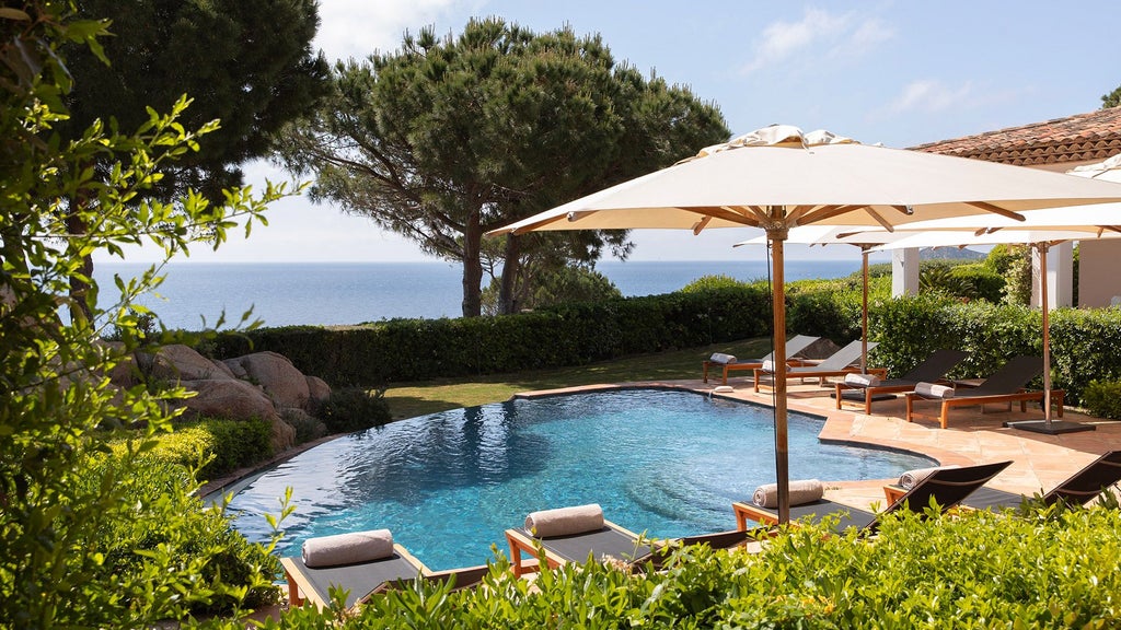 Luxurious seaside villa with white walls and blue-tiled pool overlooking Saint-Tropez coastline, surrounded by Mediterranean pine trees and elegant landscaping