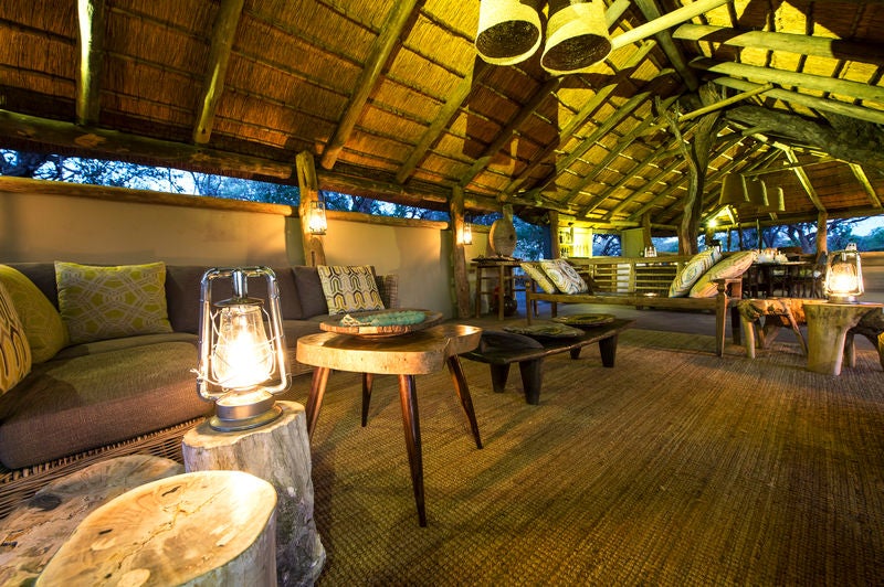 Luxurious safari tent with wooden deck overlooking pristine African wetlands at sunset, surrounded by acacia trees and tall grass