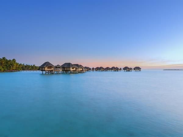 Luxurious thatched-roof overwater villa with private deck and stairs to turquoise lagoon, surrounded by vibrant coral reefs at sunset