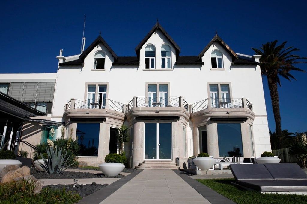 Luxurious coastal hotel with white walls and blue trim overlooking dramatic rocky shoreline and azure Atlantic waters in Cascais, Portugal