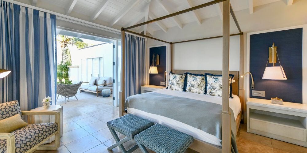 Elegant Antigua resort suite with white décor, four-poster bed, and ocean-view balcony overlooking lush tropical landscape and pristine turquoise waters