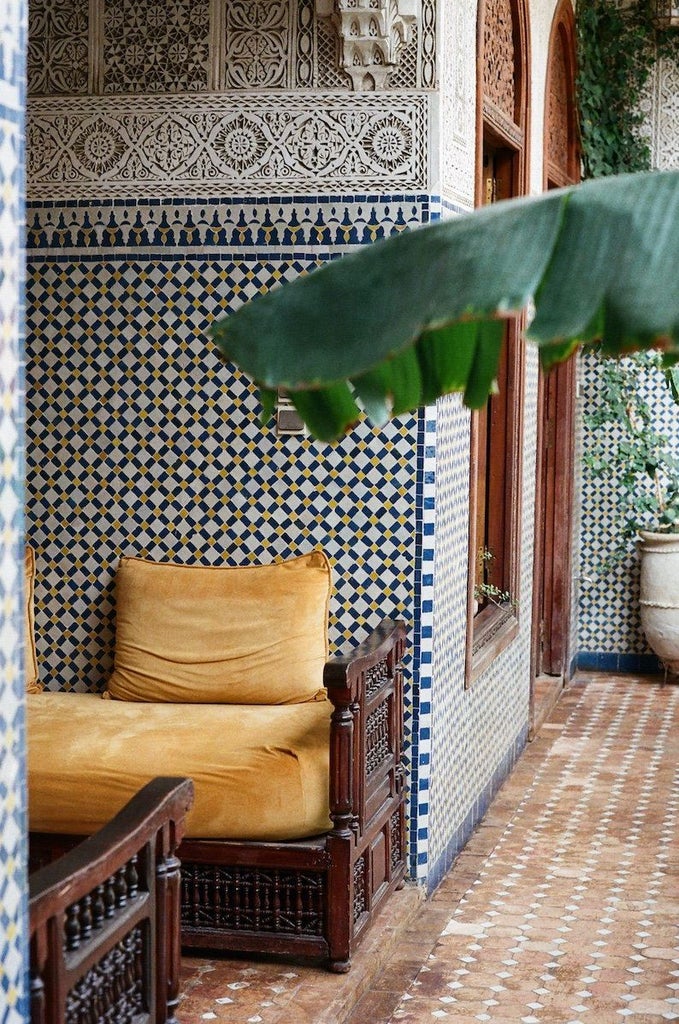 Traditional Moroccan riad courtyard with pink walls, ornate archways, lush tropical plants and mosaic-tiled fountain under blue skies