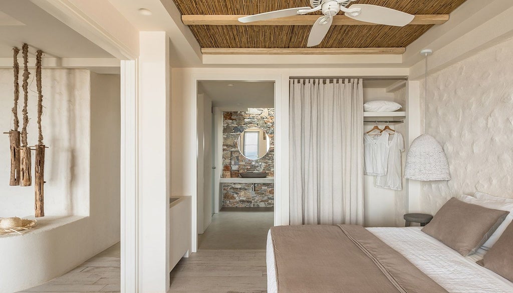 Minimalist Greek hotel room with white-draped king bed, panoramic Aegean Sea view, sleek modern furnishings, and soft natural sunlight filtering through sheer curtains