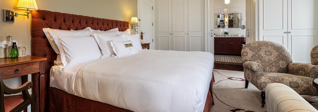 Elegant white-toned hotel room with plush king bed, ornate golden details, large windows overlooking lush Portuguese landscape at Vidago Palace Hotel