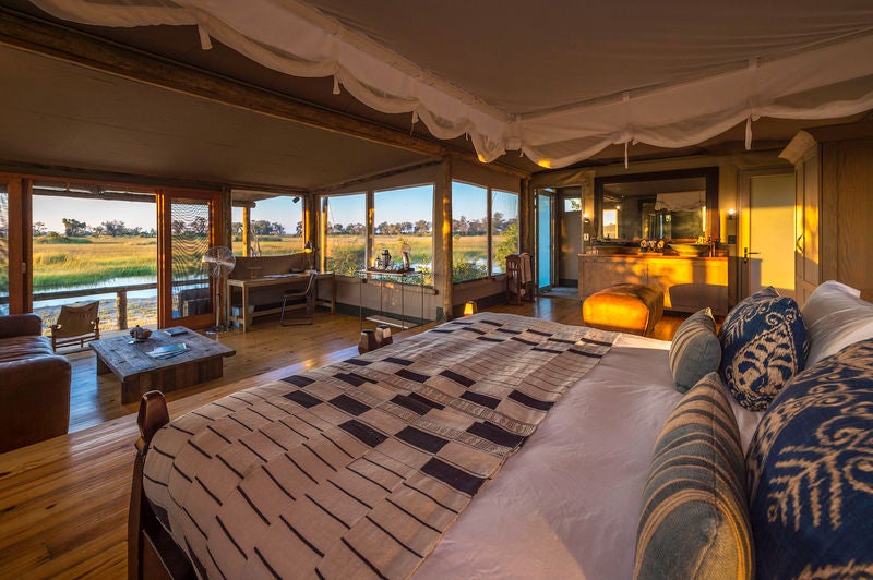 Elevated luxury safari tent with private deck overlooking Botswana wetlands, wooden walkway connecting to main lodge at sunset