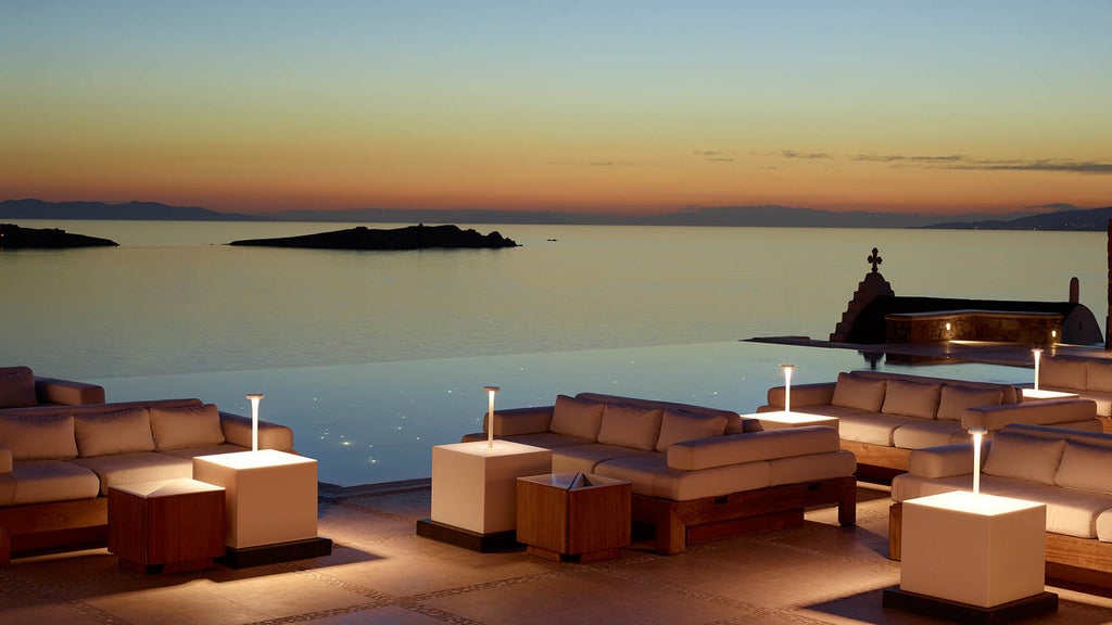 Elegant infinity pool overlooking Aegean Sea at sunset, with white lounge chairs and private dining areas set against Mykonos coastline