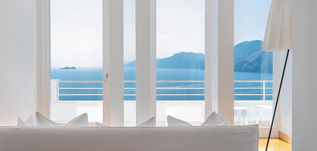 Minimalist white luxury suite overlooking Amalfi Coast, featuring elegant white furniture, expansive windows, and panoramic Mediterranean sea views at sunset