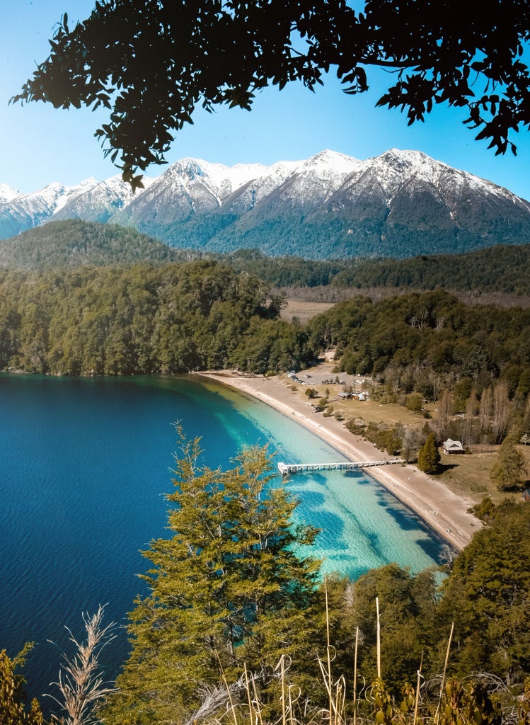 Luxurious wooden lodge perched on hillside overlooking pristine Patagonian lake, surrounded by snow-capped mountains and lush forest