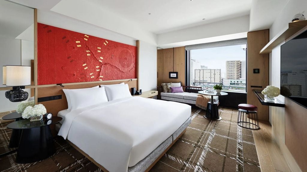 Modern hotel room with floor-to-ceiling windows offering city views, featuring a king bed, minimalist decor, and warm wood accents