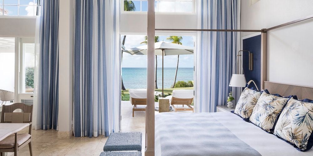 Luxurious beachside suite with white-draped four-poster bed, turquoise Caribbean waters, and pristine white sandy shoreline at Jumby Bay resort, Antigua