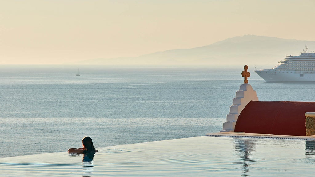 Infinity pool overlooking Mykonos bay at sunset, white loungers beside crystal-clear water, luxurious coastal architecture in background