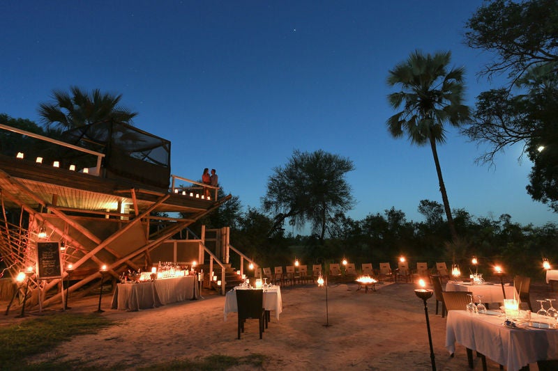 Elevated wooden walkway connects luxury safari tents overlooking African savanna at dusk, with acacia trees in the background