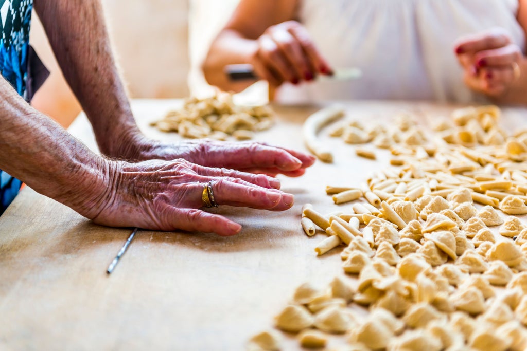 Woman in elegant kitchen showing traditional pasta-making technique on marble counter while sunlight streams through rustic window