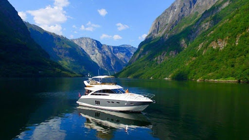 Small luxury boat cruises through narrow Norwegian fjord, towering cliffs rising on both sides, crystal-clear waters reflecting scenery