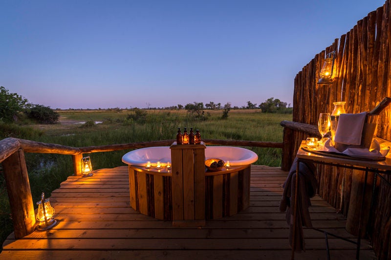 Elevated luxury safari tent with private deck overlooking Okavango Delta waters, surrounded by lush palm trees and golden grasslands