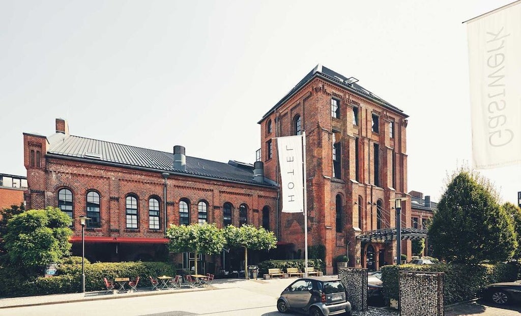 Historic red-brick industrial building converted into upscale hotel with modern glass entrance, featuring arched windows and landscaping