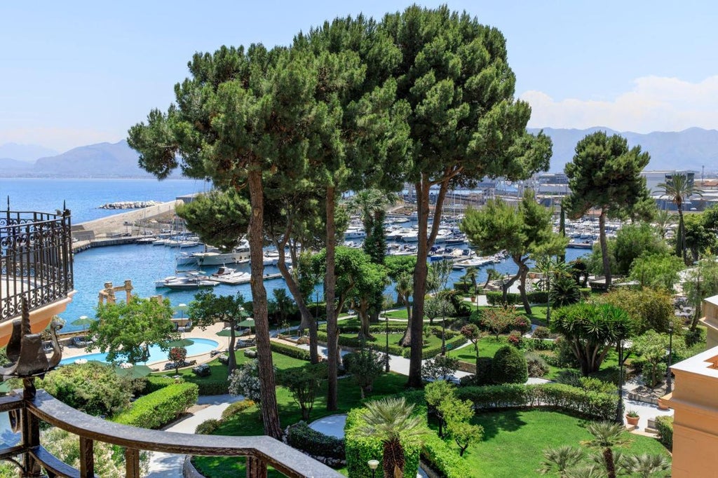 Elegant Mediterranean villa hotel with stone facade, ornate balconies, and manicured gardens overlooking Sicily's turquoise coastline